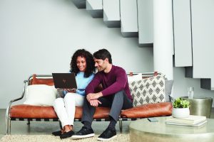 A smiling couple sit on a modern sofa, looking at a laptop together in a stylish, minimalist lounge.
