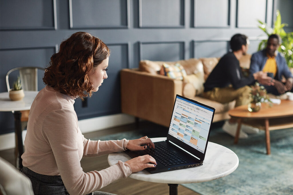 Woman using a laptop at a small table, with two people talking on a sofa in the background in a modern office setting.