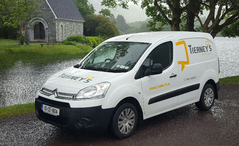 A white Tierneys delivery van is parked by a calm lake, with a stone building and trees in the background.